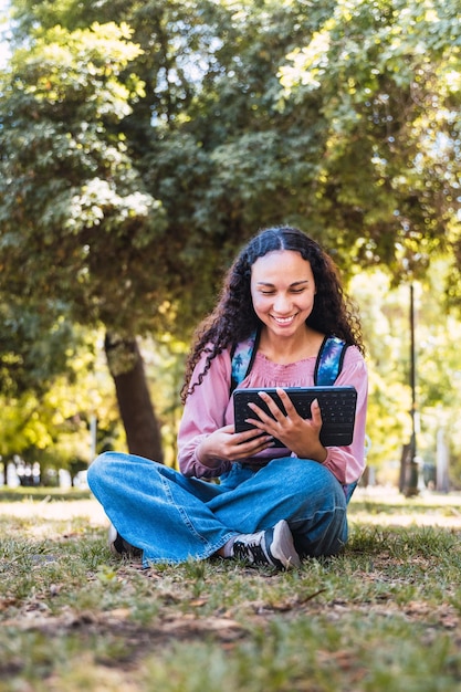 Estudante universitária negra sorrindo e usando um tablet sentado em um parque na grama