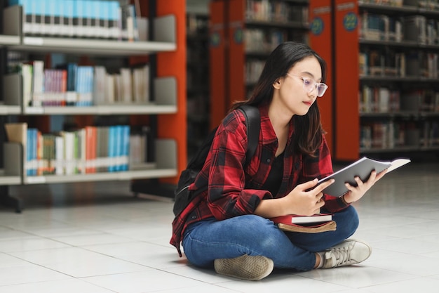 Estudante universitária usando mochila e estilo casual está sentada e lendo um livro na biblioteca.