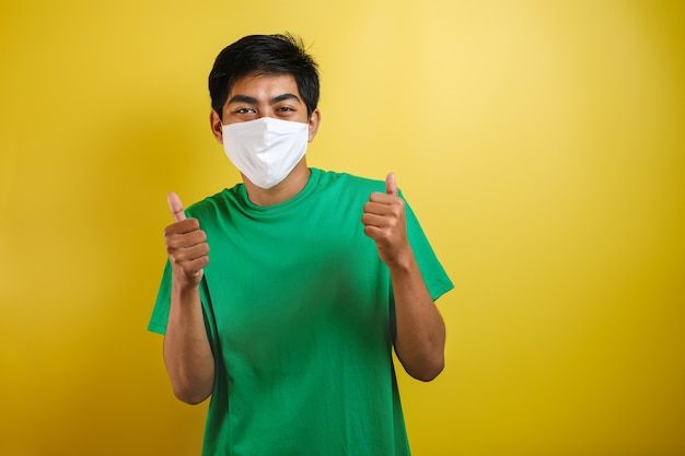 Estudante universitário asiático com o polegar para cima usando máscaras para evitar a propagação do vírus Corona contra um fundo amarelo