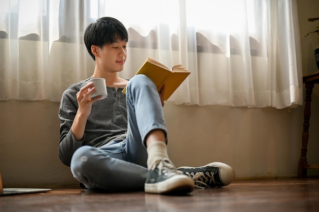 Estudante universitário asiático inteligente senta-se no chão de seu quarto lendo um livro e tomando café