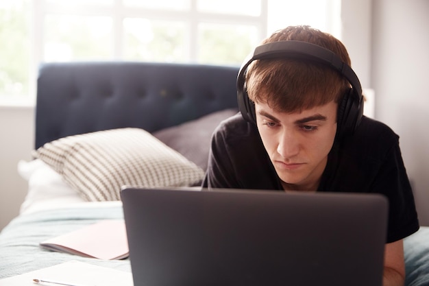 Estudante universitário masculino usando fones de ouvido deitado na cama em casa compartilhada trabalhando no laptop