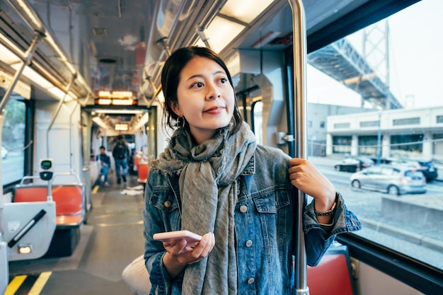 estudante universitário segurando o celular e aprendendo no vagão leve. jovem com cachecol dentro do ônibus do metrô no chão tocando a tela do celular vendo a vista urbana da cidade enquanto viaja