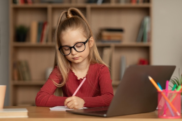 Estudante usando óculos usando laptop aprendendo on-line tomando notas dentro de casa