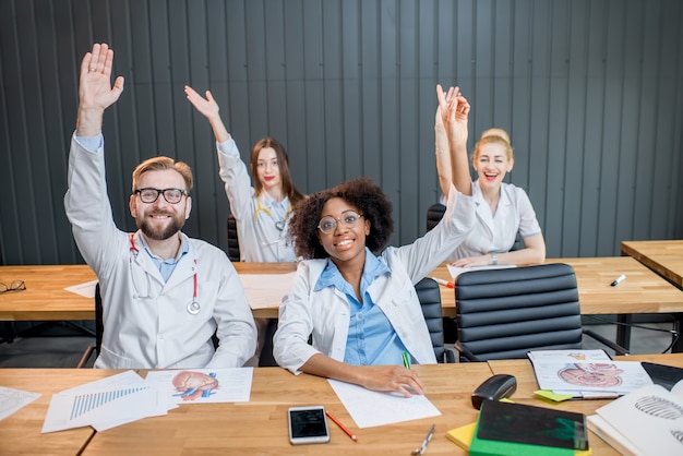 Estudantes de medicina levantando as mãos querendo responder sentados na mesa durante a aula