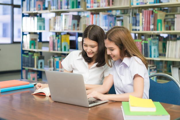 Estudantes felizes asiáticos com o laptop que trabalha e que estuda na biblioteca.