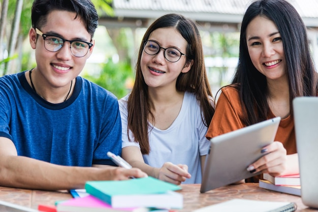 Estudantes universitários jovens sentados consultam juntos à mesa com livros, tablet e laptop.