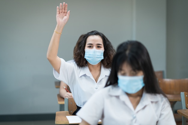 Estudantes universitários usam máscara facial em sala de aula e levantam a mão para perguntar ao professor durante a pandemia de coronavírus. Retrato de foco seletivo de estudantes universitários estudam em sala de aula.