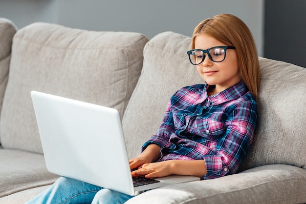 Estudar com alegria. Linda garotinha de óculos usando seu laptop enquanto está sentada no sofá em casa