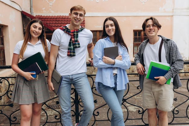 Estudar juntos é divertido conceito de construção de equipe de trabalho em equipe Quatro estudantes felizes estão sentados perto do prédio da faculdade e segurando dispositivos de livros usando um sorriso inteligente casual em um bom verão