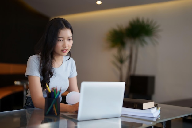 Estudiante adolescente asiática leyendo un libro y escribiendo para investigar datos en una computadora portátil para hacer tareas de asignación después de ver videos de conferencias, lecciones en línea y estudiar conocimientos educativos en un café.