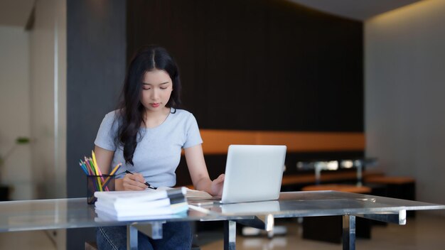Estudiante adolescente asiática leyendo un libro para tomar notas para la tarea de asignación mientras ve el video de la conferencia lección en línea en la computadora portátil para estudiar y aprender sobre el conocimiento de la educación en el café