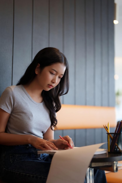 Estudiante adolescente asiática que lee información en el papeleo para tomar notas y hacer tareas para prepararse para el examen de prueba mientras estudia y aprende sobre el conocimiento educativo en el café