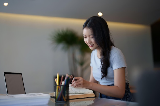 Estudiante adolescente asiática que lee un libro para escribir información del libro y hacer la tarea de la asignación después de ver la lección en línea de video en la computadora portátil y estudiar sobre el conocimiento educativo en el café