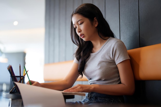 Estudiante adolescente asiática que usa lápiz de color para resaltar datos en el papeleo y tomar notas mientras lee un libro para estudiar y aprender sobre el conocimiento educativo para prepararse para el examen de prueba en un café