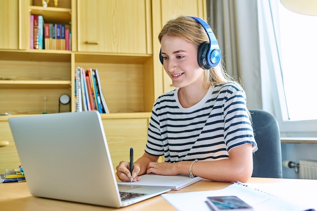 Una estudiante adolescente con auriculares sentada en un escritorio usando una computadora escribiendo en un cuaderno de estudio
