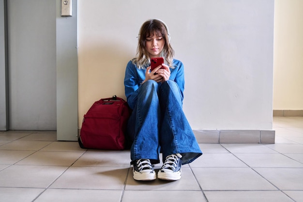 Estudiante adolescente en auriculares con teléfono inteligente de mochila sentado en el piso cerca de la pared