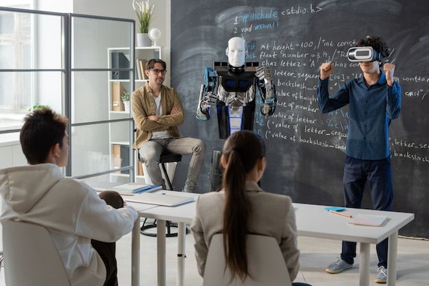 Estudiante adolescente con auriculares vr de pie junto a la pizarra delante de sus compañeros y demostrando habilidades de robot cerca