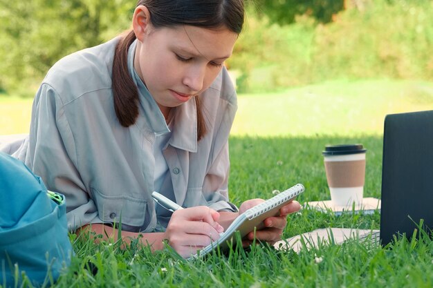 Estudiante adolescente estudiando leyendo sus libros tableta y cuaderno sentada al aire libre De vuelta a la escuela Estudiante acostada en la hierba verde usando portátil en el patio de la universidad o parque Aprendizaje a distancia