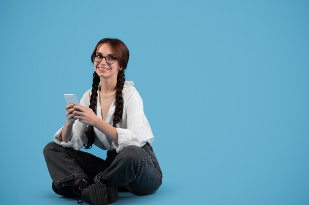 Foto una estudiante adolescente europea alegre con coletas en gafas se sienta en el suelo escribiendo en el teléfono piensa que mira el espacio vacío aislado en el fondo azul del estudio dispositivo de redes sociales para la oferta de anuncios de estudio