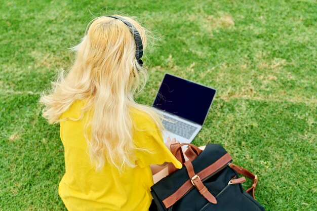 Estudiante adolescente femenina en auriculares sentado en el parque con portátil