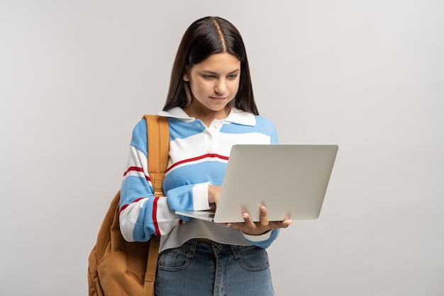 Estudiante adolescente interesada con mochila mirando la pantalla de su portátil sobre un fondo gris
