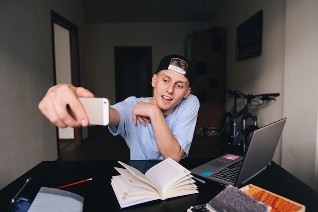 Un estudiante adolescente sonriente haciendo selfie mientras estudiaba en su casa detrás de un escritorio en la habitación. Selfies honorarios.