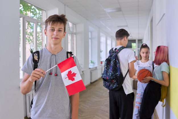 Estudiante adolescente varón con bandera de Canadá dentro de la escuela, fondo de grupo de niños. Canadá, educación y juventud, patriotismo, concepto de personas