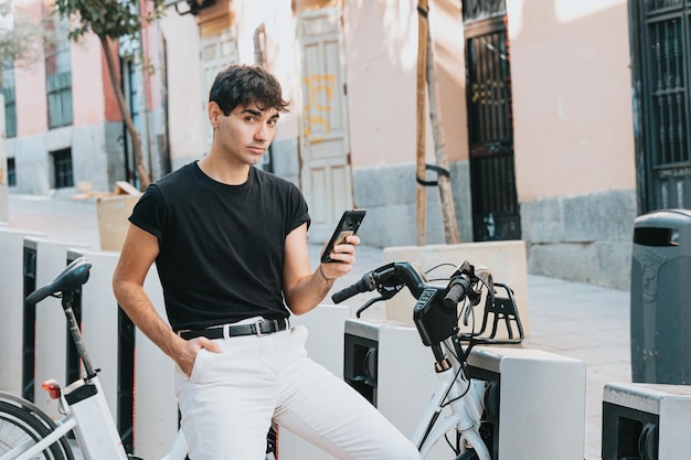 Estudiante adulto joven que alquila una bicicleta con su teléfono inteligente. Ropa moderna usando su teléfono cerca de un servicio de alquiler de bicicletas, desbloqueando bicicletas de alquiler para moverse por la ciudad, servicios de alquiler, espacio de copia