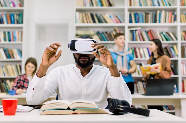 Estudiante africano masculino que usa gafas de realidad virtual en una biblioteca pública y trabaja con un libro de texto virtual para prepararse para el examen o la prueba