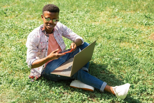 Estudiante africano sentado en el césped con un portátil y un teléfono