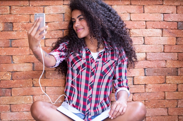 Estudiante afroamericana joven haciendo foto selfie en la cámara frontal del teléfono inteligente para compartir en redes sentado en el suelo