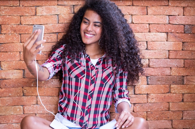 Estudiante afroamericana joven haciendo foto selfie en la cámara frontal del teléfono inteligente para compartir en redes sentado en el suelo