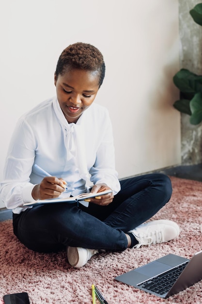 Estudiante afroamericana de pelo corto escribiendo en su cuaderno sentada en el suelo mientras usa la computadora portátil ed...