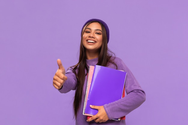 Estudiante afroamericano sonriente del adolescente de tto en un sombrero y un suéter que sostienen carpetas y que muestran el pulgar para arriba que se coloca aislado. Concepto de educacion