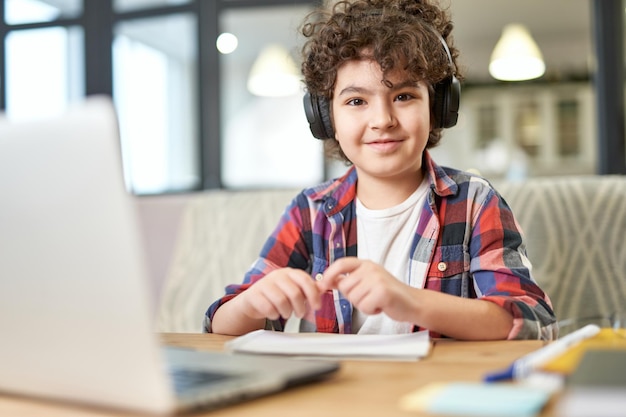 Estudiante alegre encantador niño hispano con auriculares sonriendo a la cámara mientras está sentado en el escritorio