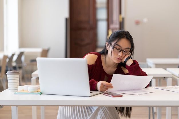 Estudiante asiática cansada y aburrida leyendo materiales impresos en papel mientras estudia en la biblioteca