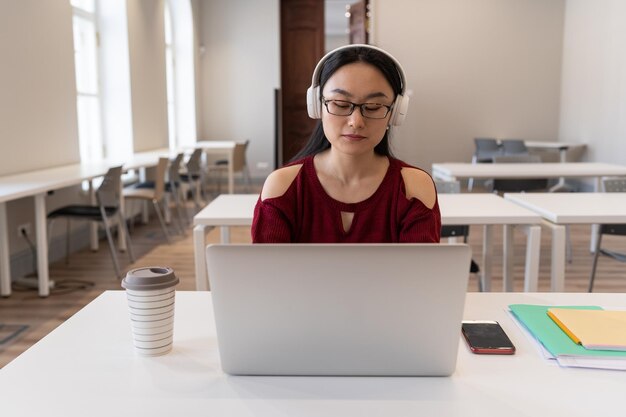 Estudiante asiática concentrada en auriculares mira el seminario web en línea o el curso de capacitación del reloj de la computadora portátil