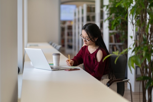 Una estudiante asiática inteligente se sienta en el escritorio, toma notas de la computadora portátil, se prepara para el examen o escribe un ensayo