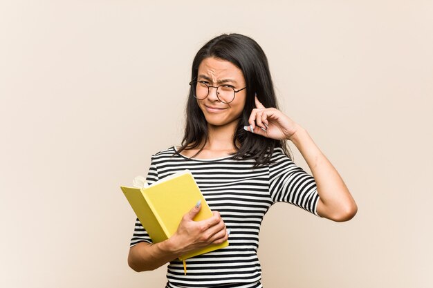 Estudiante asiática joven que sostiene un libro que cubre los oídos con las manos.