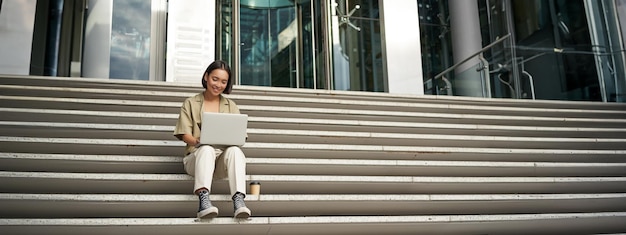 Una estudiante asiática sentada en las escaleras cerca del campus escribe en una computadora portátil y hace su tarea al aire libre