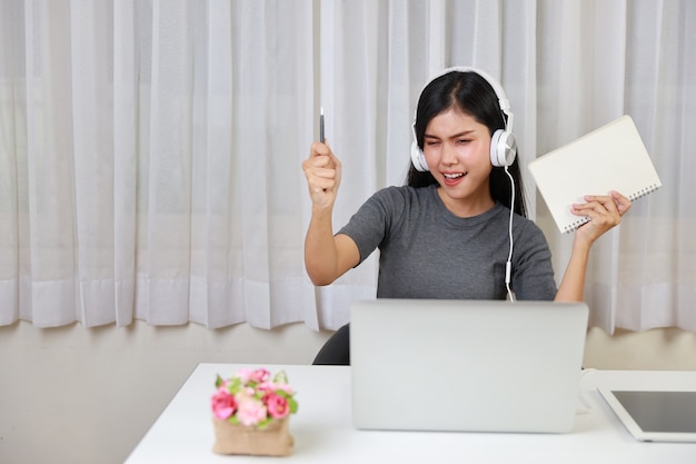 Estudiante asiática sentada a la mesa con auriculares usando la computadora y tomando nota para estudiar en línea