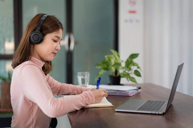 Una estudiante asiática sonriente usa auriculares inalámbricos, escribe en el cuaderno para estudiar el idioma en línea, mira y escucha al conferencista, seminario web a través de videollamadas e-learning en casa, educación a distancia