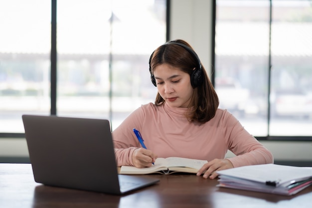 Una estudiante asiática sonriente usa auriculares inalámbricos, escribe en el cuaderno para estudiar el idioma en línea, mira y escucha al conferencista, seminario web a través de videollamadas e-learning en casa, educación a distancia