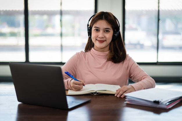 Una estudiante asiática sonriente usa auriculares inalámbricos, escribe en el cuaderno para estudiar el idioma en línea, mira y escucha al conferencista, seminario web a través de videollamadas e-learning en casa, educación a distancia