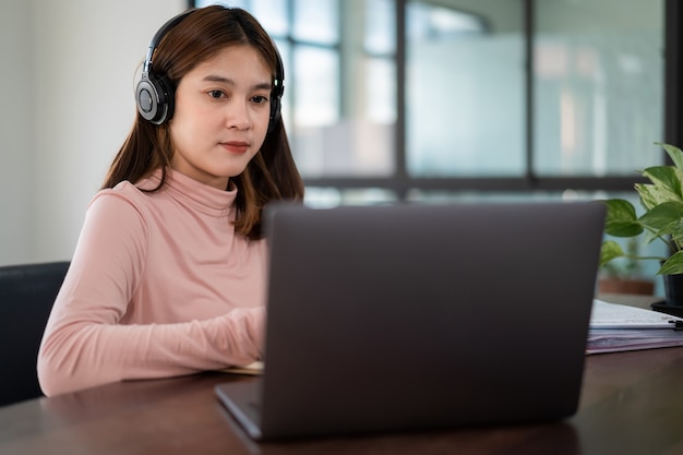 Una estudiante asiática sonriente usa auriculares inalámbricos, escribe en el cuaderno para estudiar el idioma en línea, mira y escucha al conferencista, seminario web a través de videollamadas e-learning en casa, educación a distancia