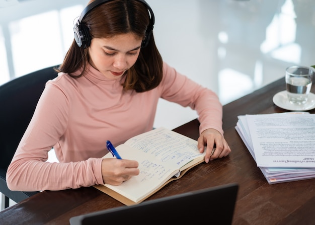 Una estudiante asiática sonriente usa auriculares inalámbricos, escribe en el cuaderno para estudiar el idioma en línea, mira y escucha al conferencista, seminario web a través de videollamadas e-learning en casa, educación a distancia