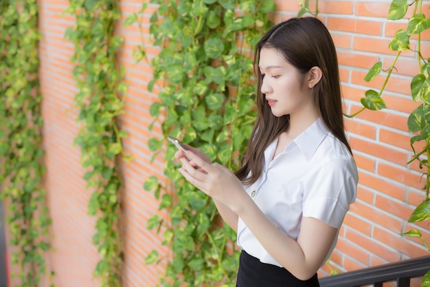 Una estudiante asiática tailandesa con uniforme sonríe alegremente mientras usa un teléfono inteligente en la universidad