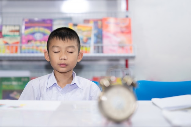 Estudiante asiático lindo del niño que medita en el reloj y el libro borrosos en el fondo blanco