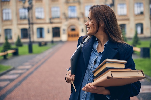 Estudiante atractiva con pila de libros y portátil sonriendo mientras está de pie cerca de la universidad sola