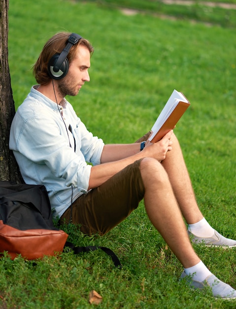 Estudiante con auriculares en el parque del campus lee un libro